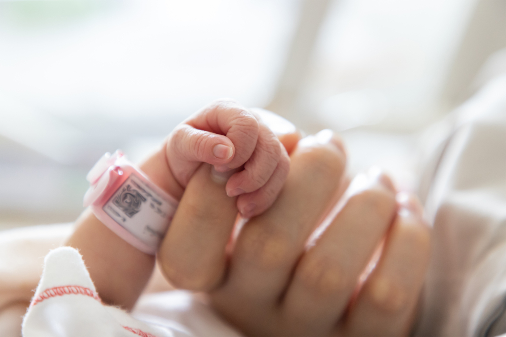 Infant hand with hospital band around wrist holding tip of index finger of an adult hand.  
